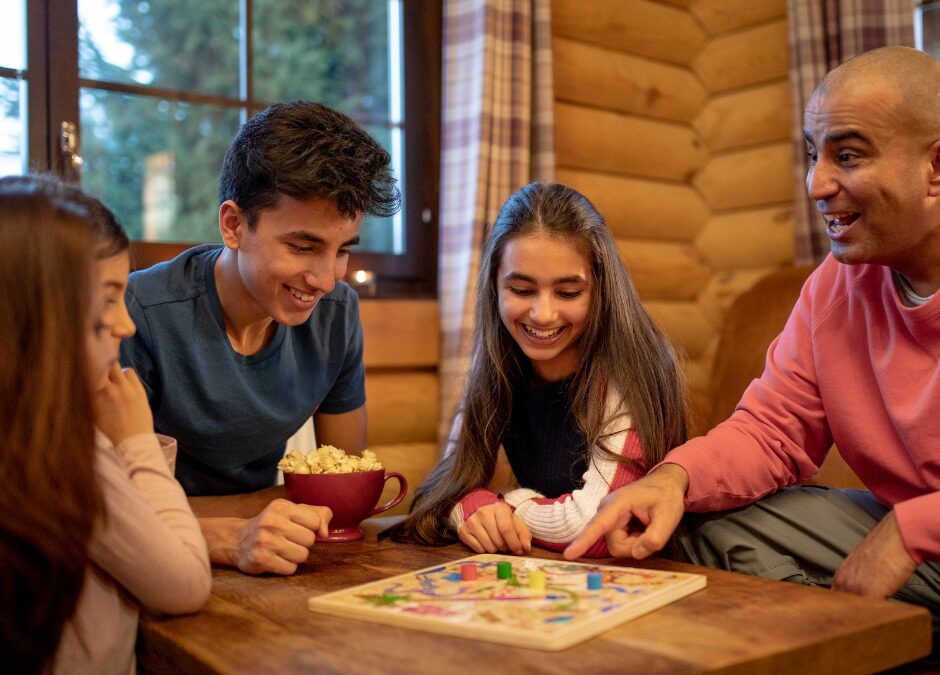 A father and three young people smile while playing a board game around a wooden table in a cozy log cabin